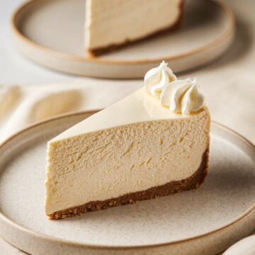 A close-up image of a slice of New York-style cheesecake with a graham cracker crust, topped with whipped cream, served on a ceramic plate.