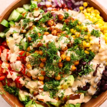 Bowl with chickpeas, corn, beans, avocado, tomatoes, cucumber, cheese, and dressing over greens and grains.